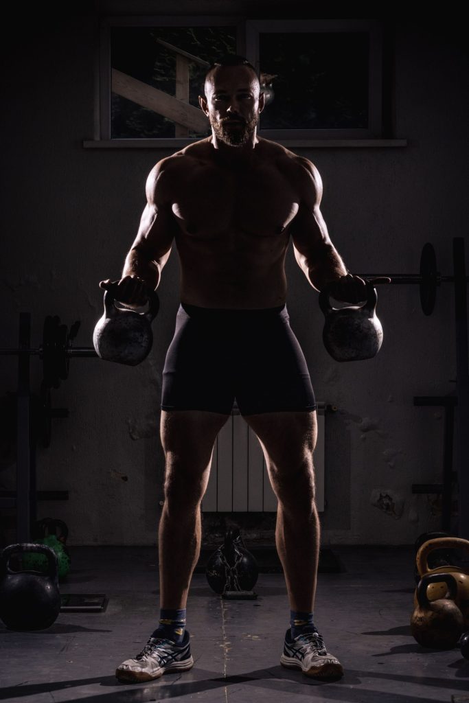 Shirtless man lifting kettlebells in dark gym, showcasing strength and fitness.